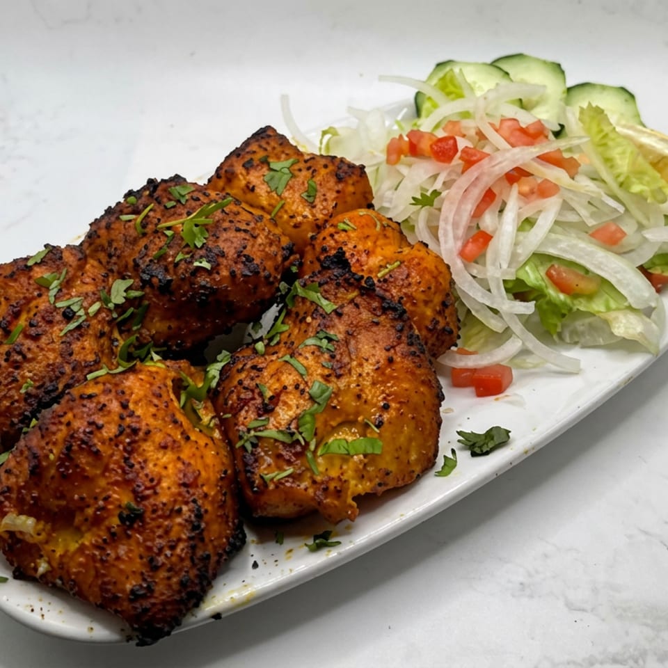Grilled spiced chicken pieces garnished with cilantro, served with lettuce, onion, tomato, cucumber, and lemon on a white platter.