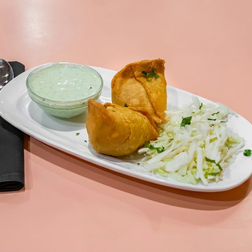 Two golden samosas with creamy green dipping sauce and shredded cabbage salad on a white plate beside silverware.