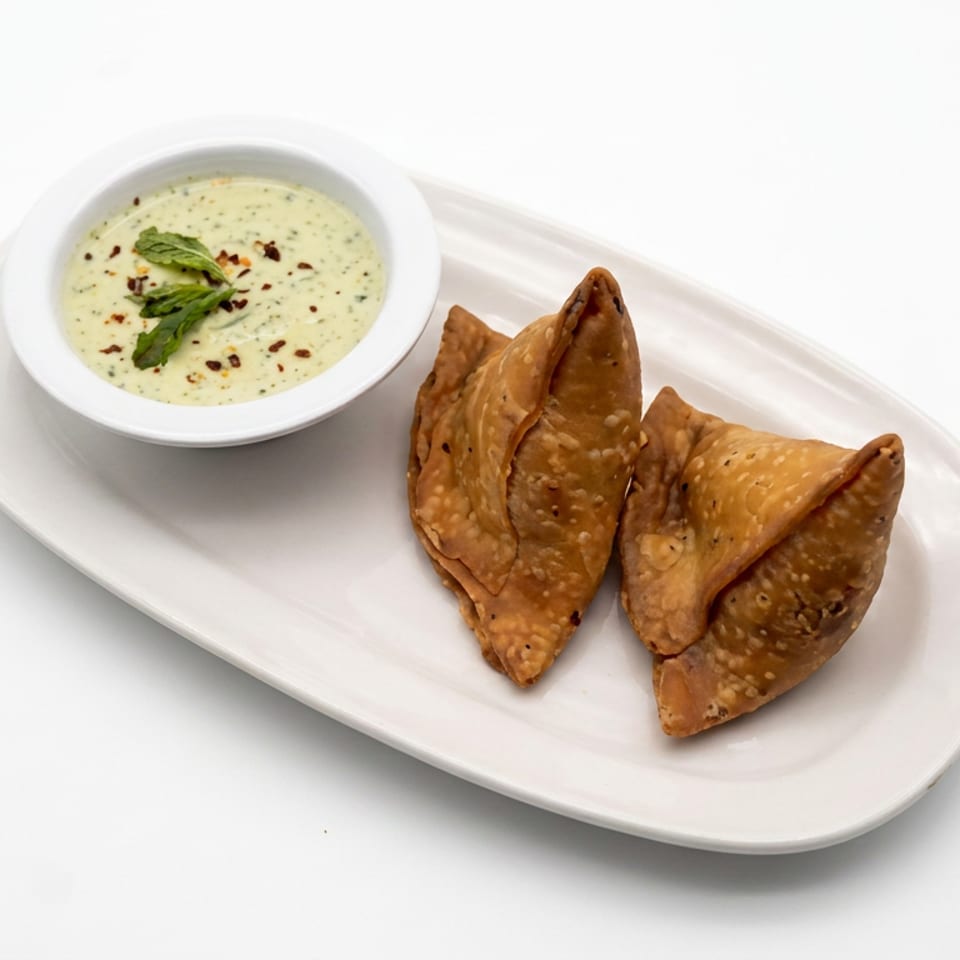Three golden fried samosas served on a white platter with creamy herb dipping sauce garnished with mint and chili flakes.